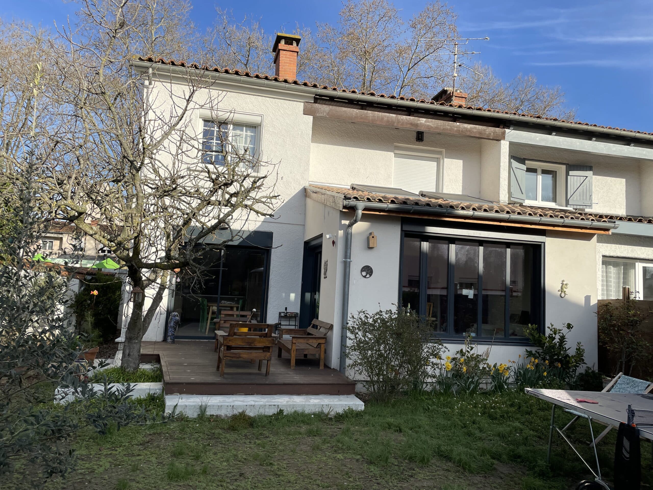 Façade arrière de maison blanche avec terrasse en bois, mobilier de jardin et arbres dénudés sous un ciel bleu clair.