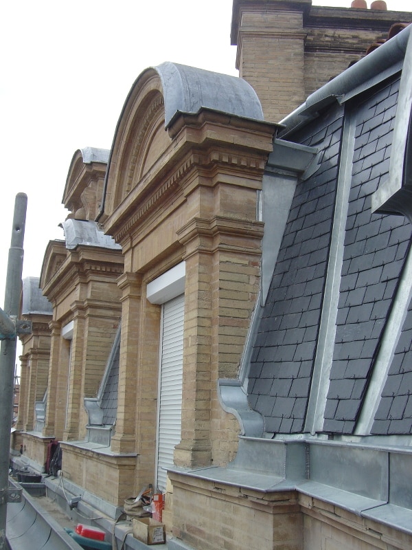Rénovation toiture mansardée : lucarnes en briques, ornements en plomb, ardoises grises et volet roulant blanc.